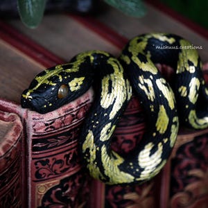 Può includere: Una statuina decorativa a forma di serpente nero e oro, realizzata a mano, arrotolata su un libro d'epoca. Il serpente presenta dettagli intricati e un design realistico. Il libro ha una copertina rossa con dettagli dorati.