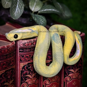 Puede incluir: Una escultura realista de una serpiente amarilla y verde descansa sobre libros rojos antiguos. La serpiente tiene un patrón texturizado y un ojo negro. Los libros tienen letras doradas y diseños decorativos. El nombre del artista, michuns.creations, es visible.