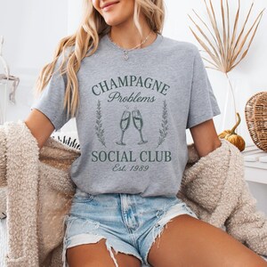 May include: A grey t-shirt with the text "Champagne Problems Social Club Est. 1989" in green. The design features two champagne flutes and floral details. The model is wearing denim shorts and a beige cardigan.