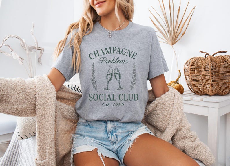 May include: A grey t-shirt with the text "Champagne Problems Social Club Est. 1989" in green. The design features two champagne flutes and floral details. The model is wearing denim shorts and a beige cardigan.