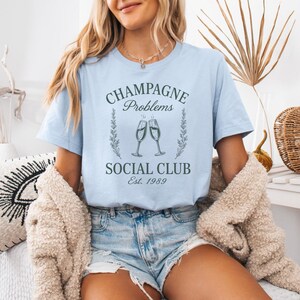 May include: Pale blue t-shirt featuring the text "CHAMPAGNE Problems SOCIAL CLUB Est. 1989" in a vintage style, with two champagne flutes. The model wears denim shorts and a beige cardigan.