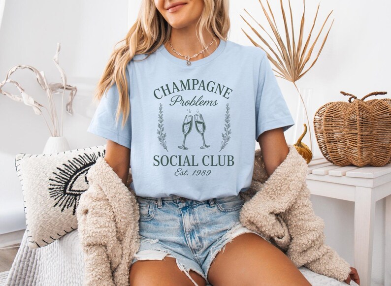May include: Pale blue t-shirt featuring the text "CHAMPAGNE Problems SOCIAL CLUB Est. 1989" in a vintage style, with two champagne flutes. The model wears denim shorts and a beige cardigan.
