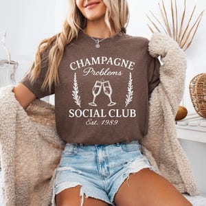 May include: A brown t-shirt with the text "CHAMPAGNE Problems SOCIAL CLUB Est. 1989" in white. The shirt features a graphic of two champagne glasses clinking. The model is wearing denim shorts and a beige cardigan.