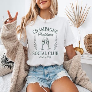 May include: White t-shirt with the text "Champagne Problems Social Club Est. 1989" in dark green. The design includes two champagne glasses. The person is wearing denim shorts and a beige cardigan.