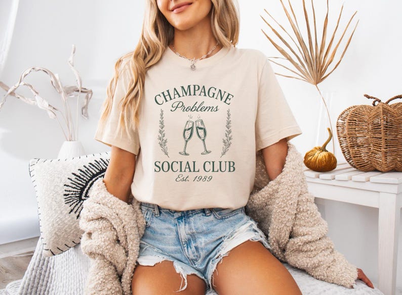 May include: A beige t-shirt featuring the text "Champagne Problems Social Club Est. 1989" in green, with champagne glasses and floral details. The shirt is worn with denim shorts and a cream cardigan.