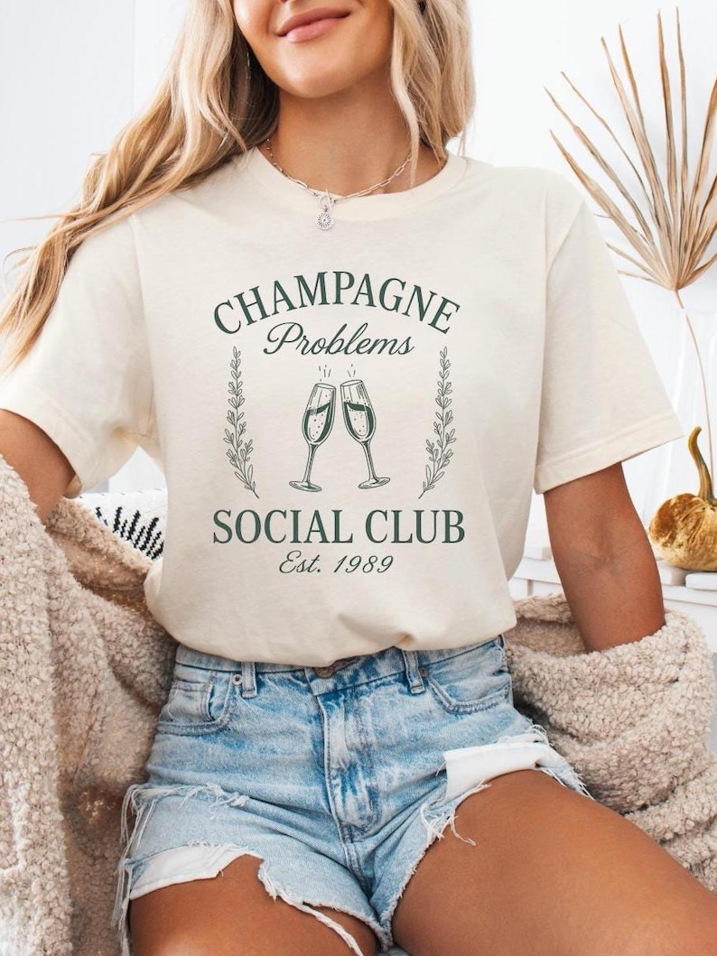May include: A cream-coloured t-shirt featuring the text "CHAMPAGNE Problems SOCIAL CLUB Est. 1989" in dark green. The design includes two champagne glasses and a laurel wreath. This is a casual wear item.