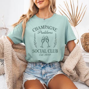 May include: A pale turquoise t-shirt featuring the text "Champagne Problems Social Club Est. 1989" in dark green. The design includes two champagne glasses and decorative leaves. The shirt is worn with denim shorts.