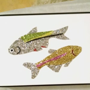 Könnte beinhalten: Zwei glitzernde Fischkunstwerke auf weißem Hintergrund, in Schwarz gerahmt. Ein Fisch ist silber mit einem grünen Streifen, der andere goldfarben mit einem rosa Streifen. Die Fische haben einfache, cartoonartige Merkmale.