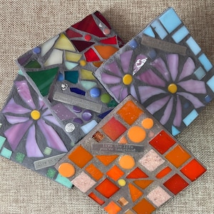 May include: Four square mosaic coasters with inspirational quotes. Each coaster features a unique design with colorful glass pieces. One coaster has a floral design, and another has an orange geometric pattern. The coasters are on a neutral background.