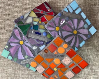 May include: Four square mosaic coasters with inspirational quotes. Each coaster features a unique design with colorful glass pieces. One coaster has a floral design, and another has an orange geometric pattern. The coasters are on a neutral background.
