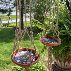 May include: Two hanging planters made of terracotta with mosaic tile bottoms. The planters are suspended by macrame cords and are hanging in a garden setting.