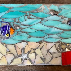 May include: A rectangular mosaic art piece featuring a sea scene with light blue and turquoise glass shards representing water. A blue and white striped fish with an orange face, a starfish, and two small metallic fish are embedded in the design.