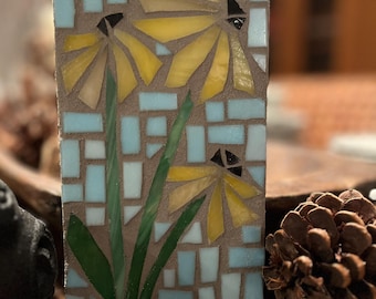 May include: A rectangular mosaic art piece featuring three yellow flowers with black centers and green stems against a light blue and brown background. The mosaic is made of small, colorful tiles, creating a textured surface.