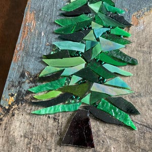 May include: A mosaic art piece featuring a stylized Christmas tree. The tree is composed of various shades of green glass pieces, with a dark brown base. The mosaic is set against a weathered, gray-toned wooden background.