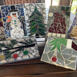 May include: A collection of mosaic art pieces featuring holiday-themed designs. The mosaics depict a snowman, Christmas tree, Santa hat, and ornaments. The pieces are made of colorful tiles and set on a wooden surface.