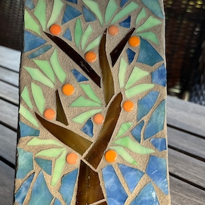 May include: A mosaic art piece depicting a tree with green leaves and orange fruit. The tree is set against a background of blue and green mosaic tiles. The piece is mounted on a metal stand.