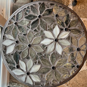 May include: Round mosaic table top with a floral design. The flowers are made of glass in shades of white, green, and gray, with black centers. The mosaic is set in a gray grout, and the table has a metal rim.