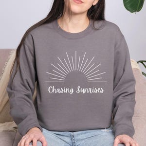 May include: A charcoal grey sweatshirt with a white graphic of a stylised sun with the text "Chasing Sunrises" underneath.