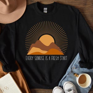 May include: Black sweatshirt featuring a graphic of a sunrise over mountains and the text "Every sunrise is a fresh start".