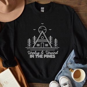 Pode incluir: Camisola de moletom preta com um design gráfico branco de uma cabana na floresta com o texto "Unplug & Unwind in the Pines".