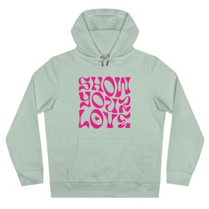 Può includere: Felpa con cappuccio verde chiaro con tasca frontale e cappuccio con coulisse. La parte anteriore della felpa presenta la scritta "SHOW YOUR LOVE" in rosa, con un font in stile retrò. La felpa è realizzata in materiale morbido.