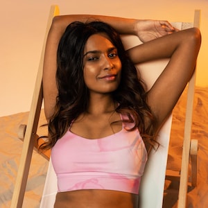 Op de afbeelding: Een vrouw draagt een roze sportbeha met een bloemenpatroon en ontspant in een houten strandstoel.