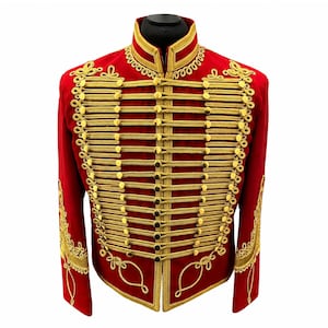 Puede incluir: Una chaqueta roja con detalles de cordón trenzado dorado en la parte delantera y las mangas. La prenda incluye un cuello alto y múltiples botones dorados dispuestos en filas horizontales, con intrincados bordados dorados.