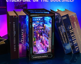 May include: A miniature cyberpunk diorama with a detailed street scene, illuminated by blue and purple lights, is displayed between several books. The books have titles like "A-Declining Steel Town" and "Alliance". The diorama has the text "Cyberworldworld".