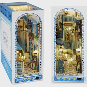 May include: A miniature diorama of a seaside village scene, featuring blue and white buildings, cobblestone streets, and small tables and chairs. The diorama is housed in a box with the text "Sea Breeze".