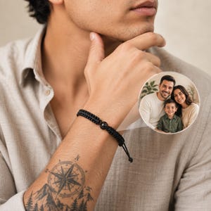 Puede incluir: Una pulsera de cordón trenzado negro con un dije circular central. El dije contiene una pequeña fotografía circular a todo color de una familia compuesta por un hombre, una mujer y un niño pequeño, todos sonriendo en un entorno interior luminoso.