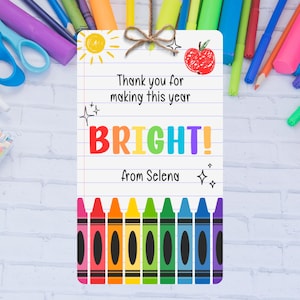 May include: A vibrant bookmark featuring a lined paper design. The text says "Thank you for making this year BRIGHT! from Selena." A row of crayons in various colours is at the bottom, with a sun and apple illustration at the top.