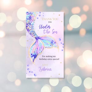 Può includere: Biglietto di ringraziamento a forma di coda di sirena viola e blu con accenti di glitter dorati. Il biglietto dice "Grazie da Under the Sea" e "Per aver reso il mio compleanno speciale! Con affetto, Sabrina".