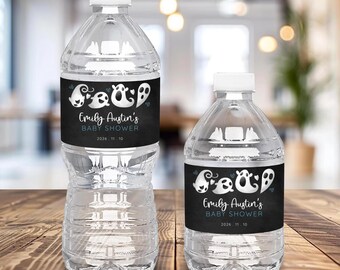 Puede incluir: Dos botellas de agua de plástico transparente con tapas blancas. Cada botella tiene una etiqueta negra con ilustraciones de fantasmas blancos y el texto "Emily Austin's BABY SHOWER 2026. 11. 10". Las botellas están sobre una superficie de madera.
