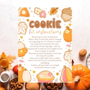 May include: A decorative sign with the text "COOKIE kit instructions" in brown. The sign features illustrations of cookies, pumpkins, and autumn leaves in shades of orange, pink, and white. Instructions are provided for cookie decorating.
