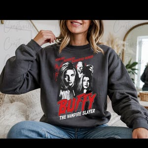 Puede incluir: Sudadera gris oscuro con un gráfico de la serie de televisión Buffy the Vampire Slayer. El gráfico incluye imágenes de personajes y el título del programa en letras rojas. El texto "Demons, Darkness, Dangerous Women" está encima de las imágenes.