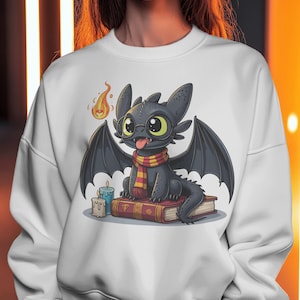 Op de afbeelding: Witte sweatshirt met een cartoon draak met een bril en een sjaal. De zwarte draak met gele ogen en een rood-gouden sjaal zit op boeken. Een kaars en een vlam zijn ook aanwezig.