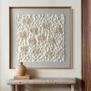 Op de afbeelding: Een ingelijst wandkunstwerk met een wit, textuurrijk ontwerp van bloemen gemaakt van kleine, ronde, witte objecten. De kunst hangt aan een witte muur boven een houten tafel met een vaas en boeken.