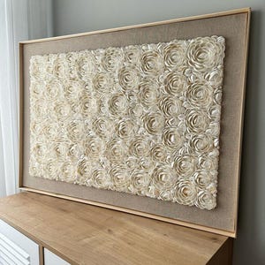 Op de afbeelding: Een groot ingelijst wandkunstwerk met een beige achtergrond en een textuurpatroon van witte schelpbloemen.