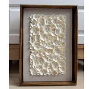Op de afbeelding: Een ingelijst wandkunstwerk met een textuurontwerp van witte bloemblaadjes. De bloemblaadjes zijn gerangschikt in een dicht, overlappend patroon, waardoor een 3D-effect ontstaat. De lijst is gemaakt van hout en heeft een natuurlijke afwerking.