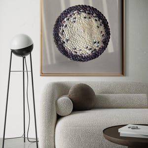 Può includere: Un'opera d'arte incorniciata che presenta un design circolare realizzato con piccole conchiglie bianche e viola. L'opera d'arte è appesa a una parete bianca sopra un divano bianco con un cuscino marrone.