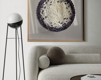 May include: A framed artwork featuring a circular design made of small, white and purple seashells. The artwork is hanging on a white wall above a white couch with a brown pillow.
