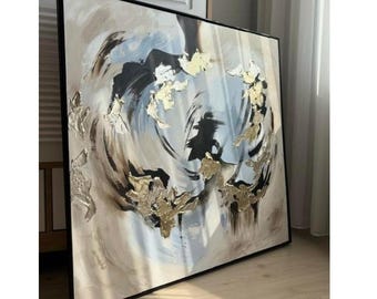 May include: Abstract painting with a black frame. The artwork features swirling brushstrokes in shades of white, black, and brown, with accents of light blue. Gold leaf embellishments are scattered throughout the composition, adding texture and visual interest.