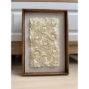 May include: A framed artwork featuring a rectangular arrangement of cream-coloured, three-dimensional flowers. The flowers are set against a neutral fabric background within a dark brown wooden frame. The artwork is displayed on a light-coloured surface.