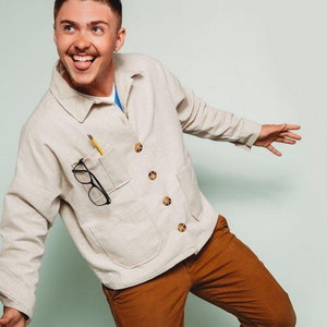 May include: A man wearing a light beige button-up shirt with a pencil and glasses in the pocket. He is wearing brown pants and white sneakers with green accents.