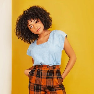 Peut inclure: Une femme portant un haut bleu clair à manches courtes et un pantalon à carreaux orange et noir. Elle se tient devant un mur jaune et un mur blanc.