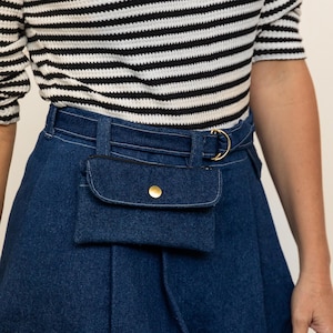 Puede incluir: Una falda de mezclilla azul con un bolsillo grande en la parte delantera. La falda tiene un cinturón de cuero marrón con una hebilla dorada y un anillo dorado.