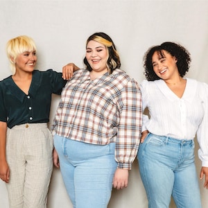 Könnte beinhalten: Drei Personen stehen zusammen und lächeln. Die Person in der Mitte trägt ein kariertes Hemd und eine blaue Jeans. Die Person links trägt ein dunkelgrünes Hemd und eine karierte Hose. Die Person rechts trägt ein weißes Hemd und eine blaue Jeans.