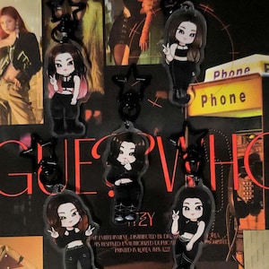 May include: Keychains featuring cartoon illustrations of a person in black outfits. The keychains are hanging against a backdrop with the words "GUESS WHO" and "Phone" visible. The keychains have black star-shaped attachments.