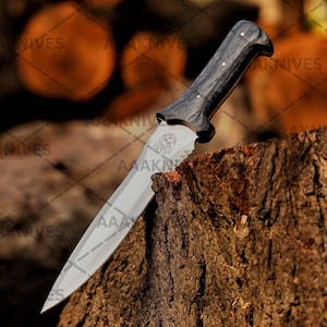 May include: A knife with a polished steel blade and a dark wooden handle. The handle has a curved design and is secured with small silver rivets. The blade has a decorative emblem near the handle. The knife is resting on a tree stump.