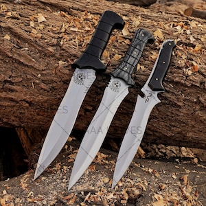 May include: Three daggers with silver blades and black handles are displayed on a wooden surface. The handles have different designs, including a curved shape and a textured grip. Each blade has a decorative emblem near the handle.